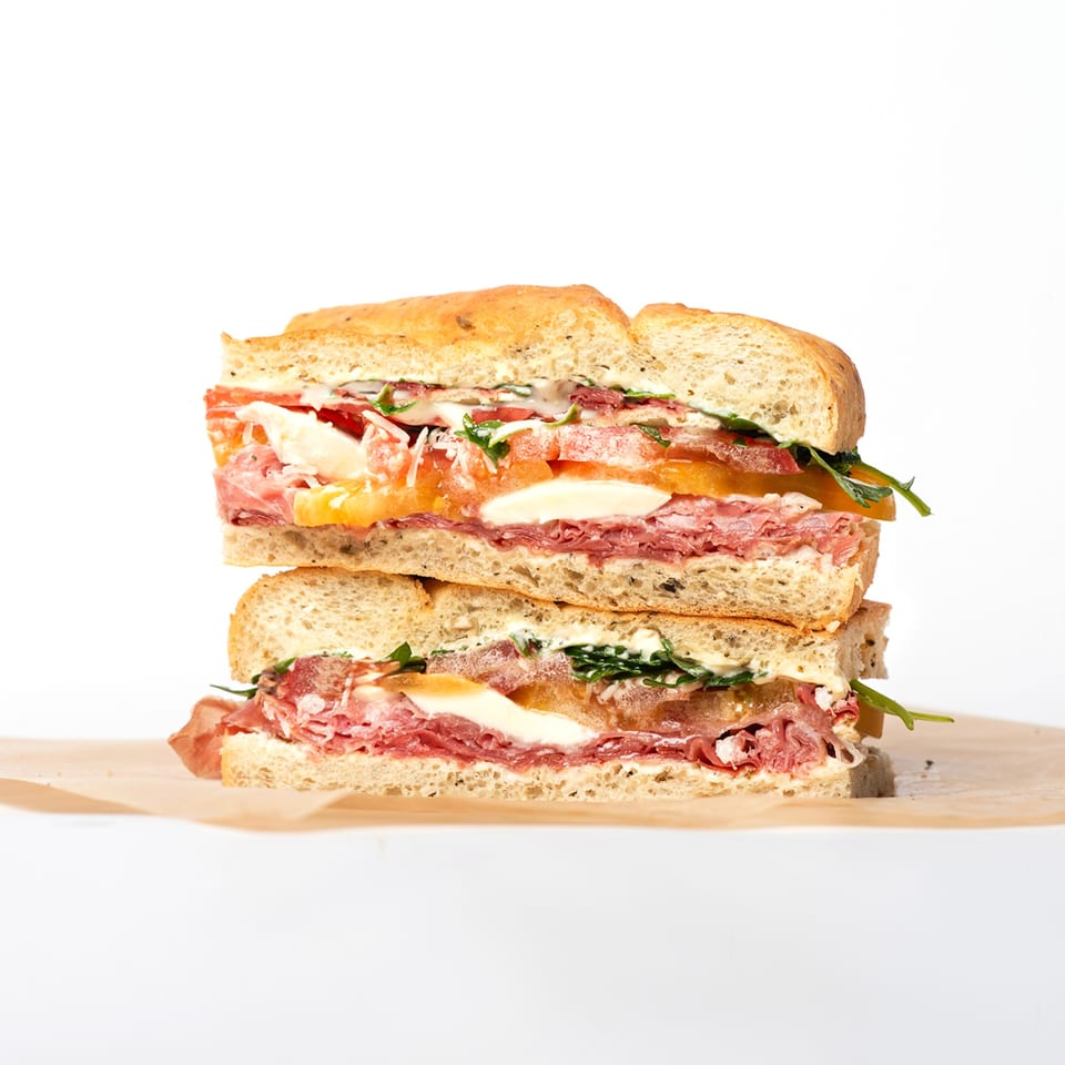 A Deliche sandwich, cut in half and stacked, revealing layers of meat, cheese, tomatoes, and greens.