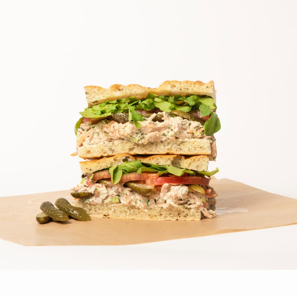 A large Deliche tuna salad sandwich, cut in half and stacked, with pickles on the side.