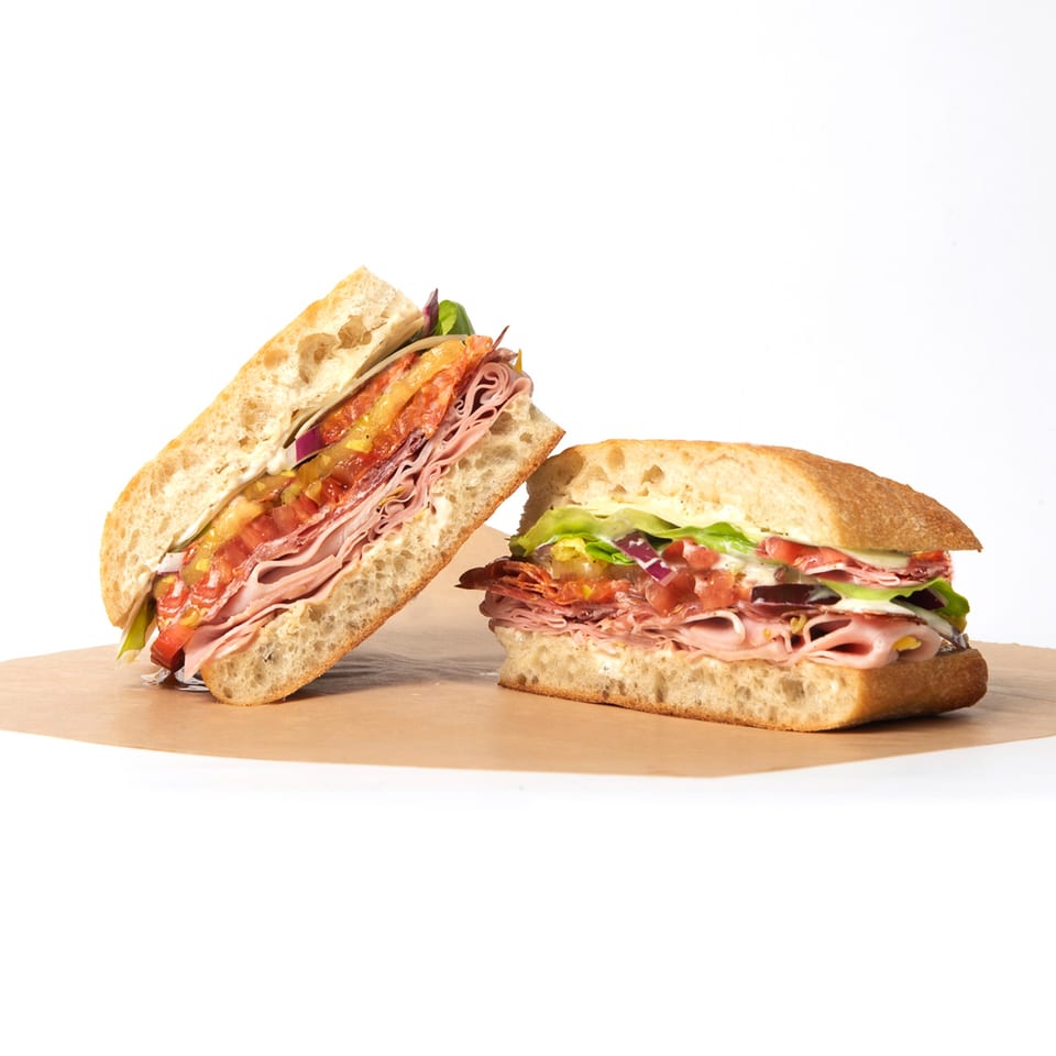 Two Deliche sandwiches, one cut in half, revealing layers of meat, cheese, and vegetables.